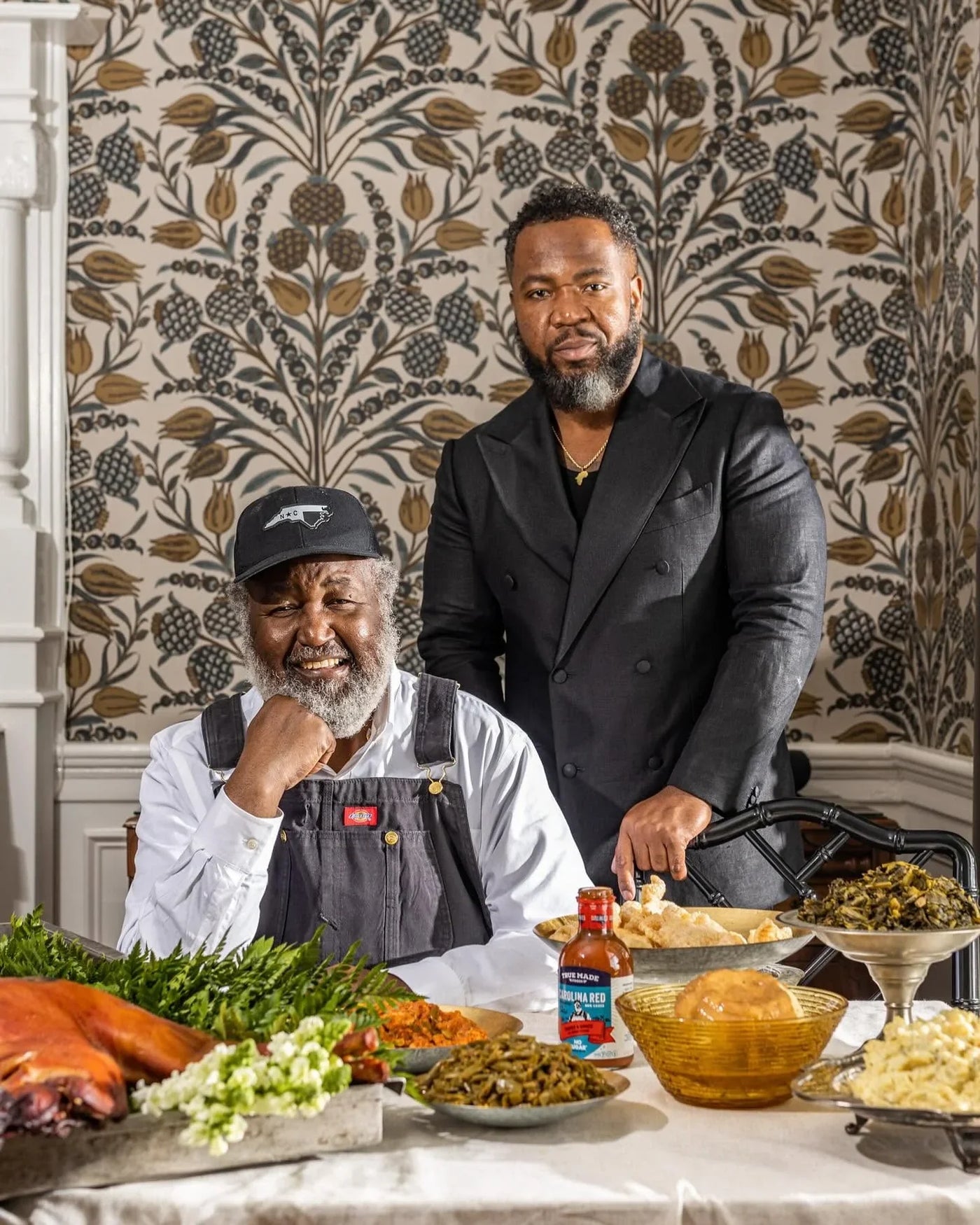 Pitmasters Ed and Ryan Mitchell seated at a table with traditional southern barbecue and True Made Foods BBQ sauce.