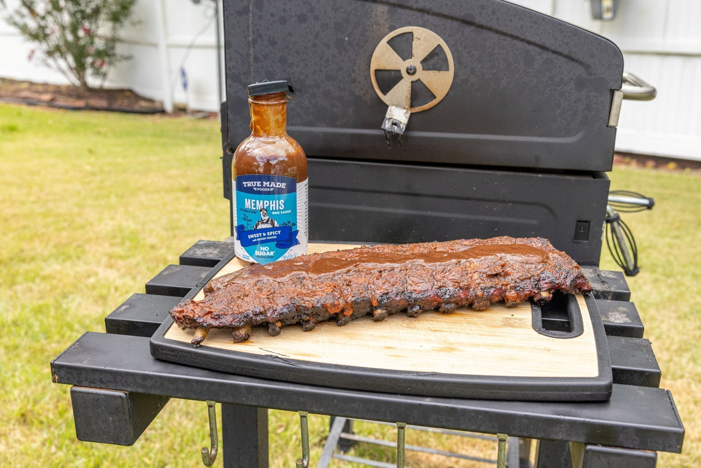 Bottle of True Made Foods Memphis BBQ sauce displayed beside grilled ribs on a cutting board near an outdoor barbecue grill.