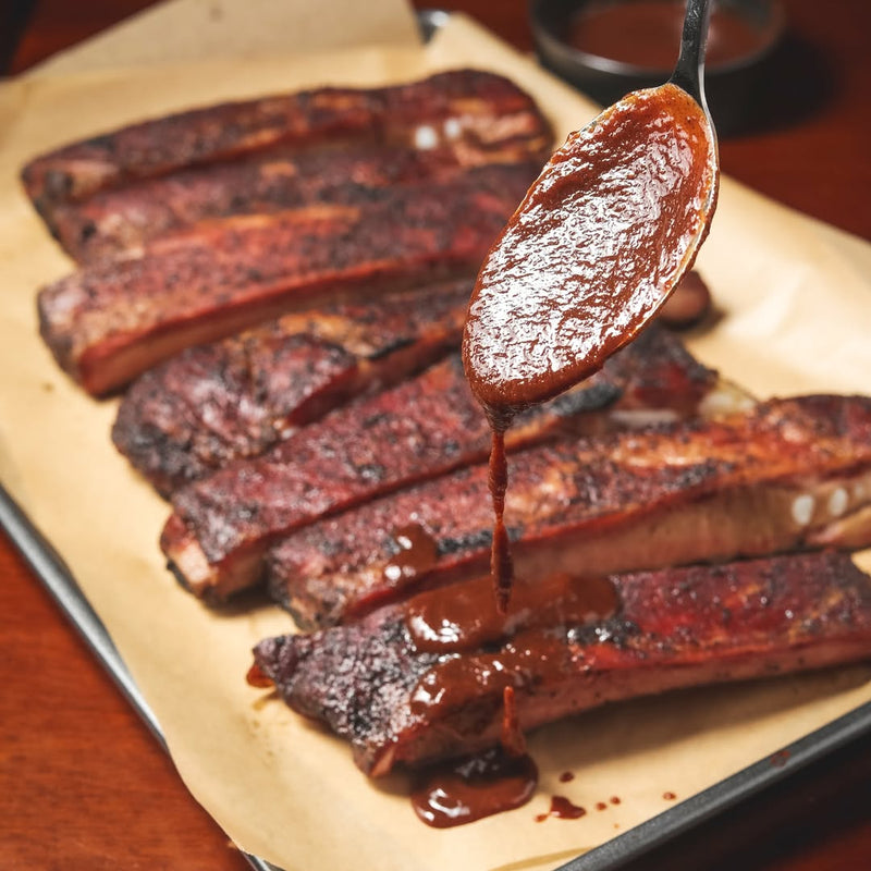 A spoon pouring True Made Foods Tomato BBQ sauce over barbecue pork ribs on a tray.