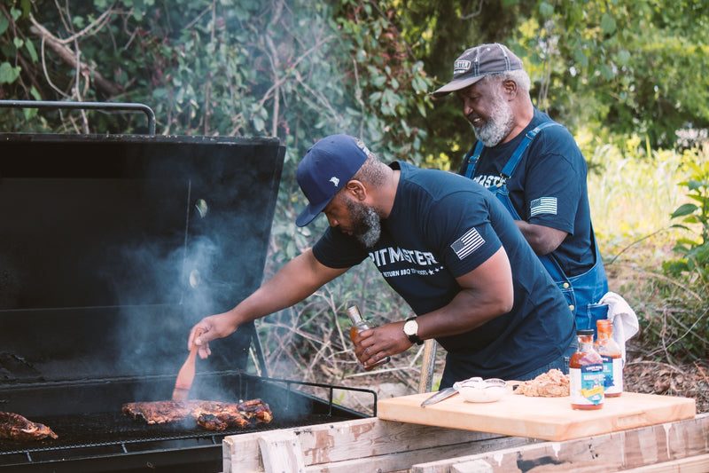 Ed Mitchell and Ryan Mitchell barbecuing in their yard, brushing True Made Foods sugar-free BBQ sauce onto meat.