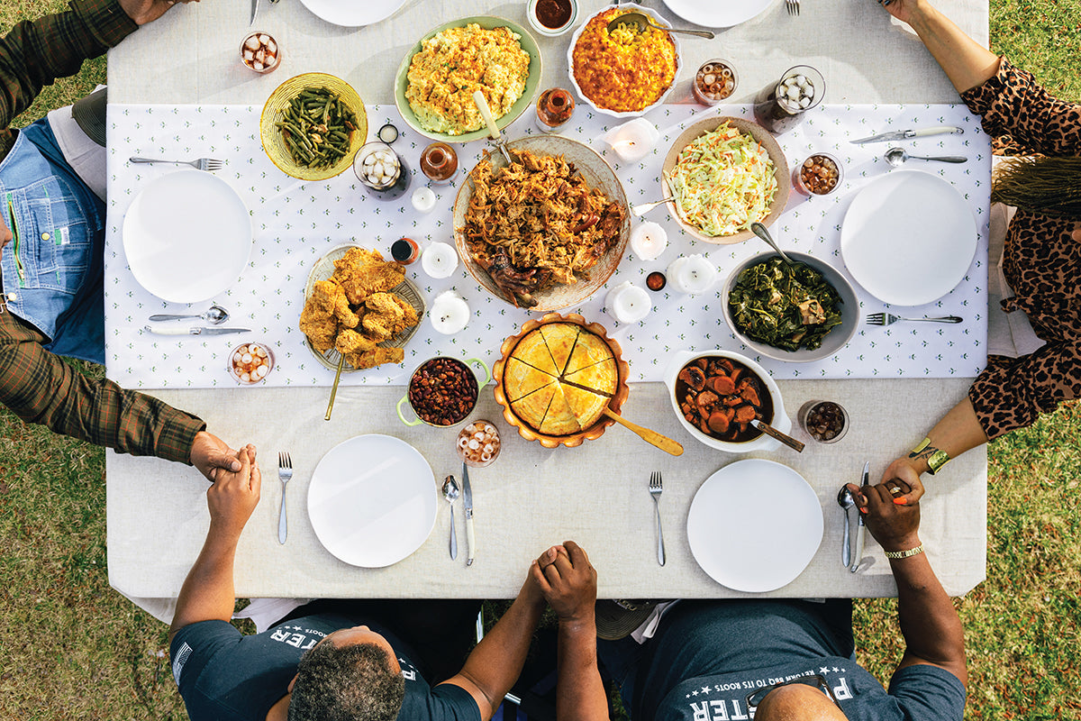 Friends and family holding hands around a Christmas table featuring BBQ mains, vegetable sides, and True Made Foods sauces.