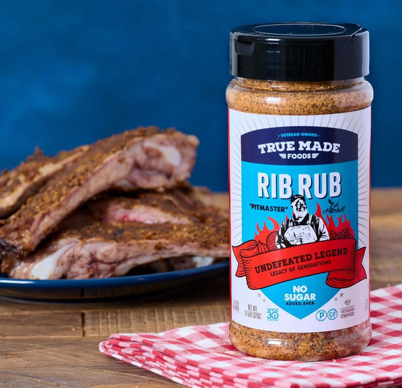 Jar of True Made Foods No Added Sugar Rib Rub beside a plate of seasoned barbecue ribs ready for grilling.