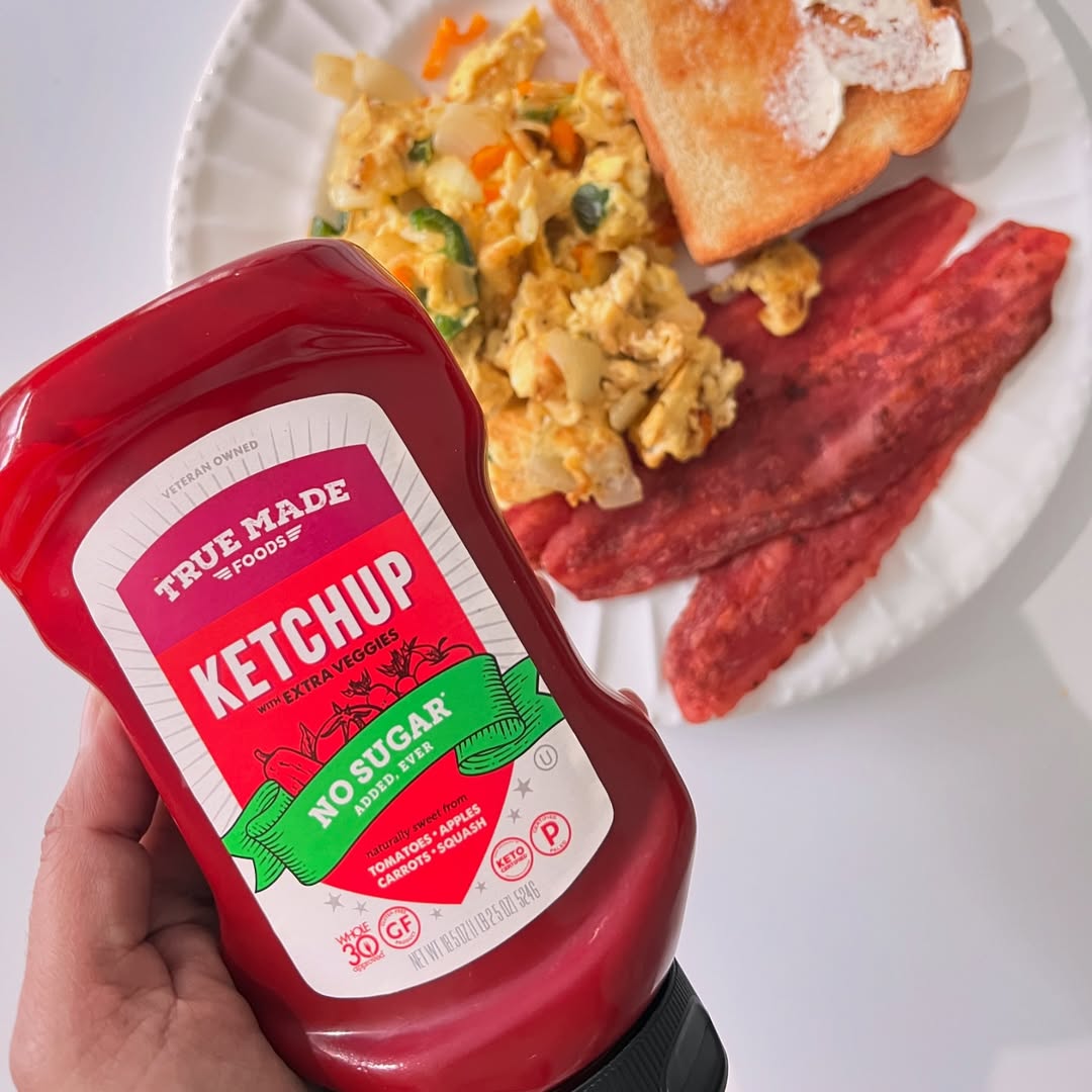 Hand holding a bottle of True Made Foods no-sugar ketchup in front of a breakfast plate with eggs, bacon, and toast.