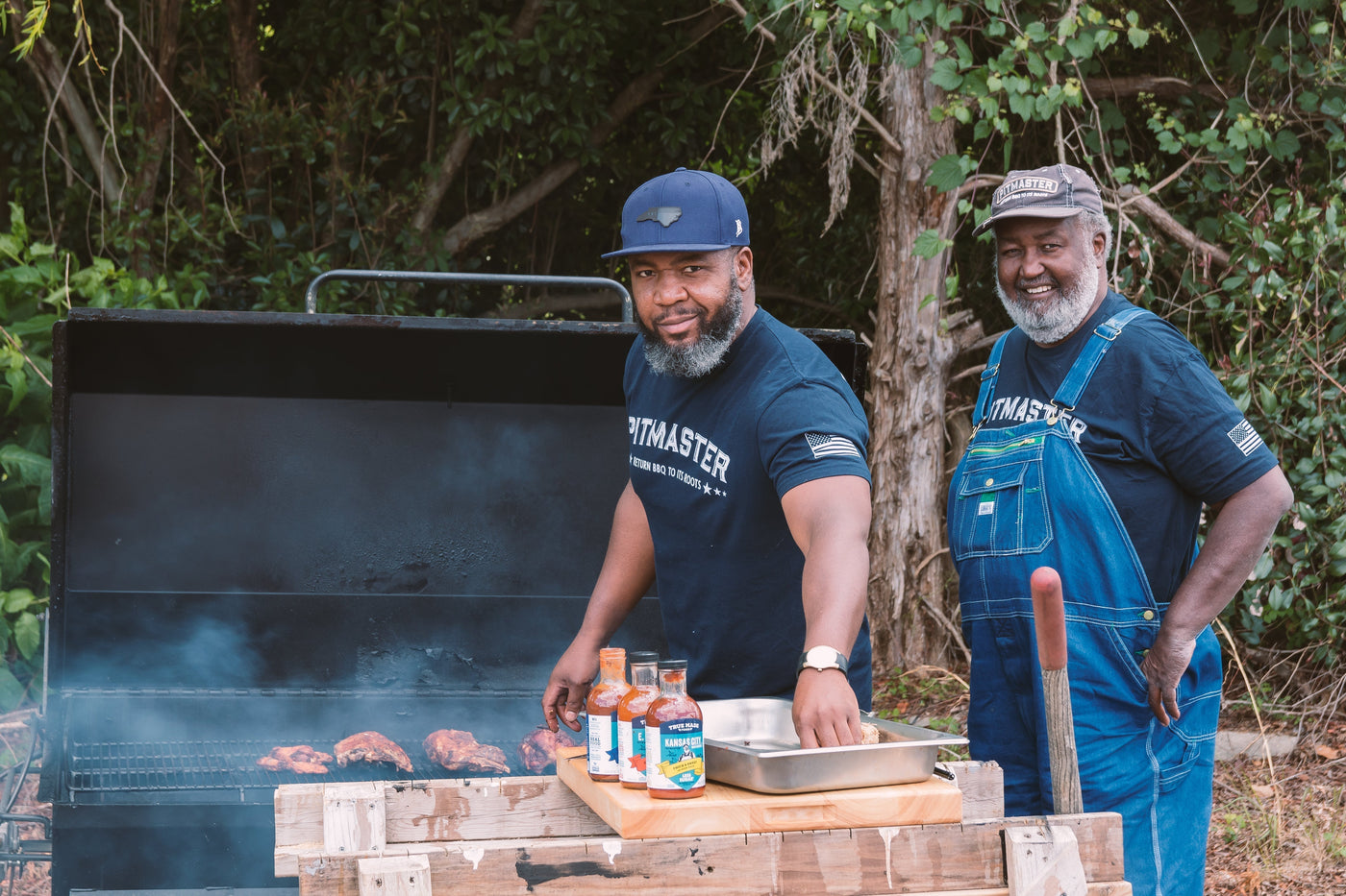 Two pitmasters, Ed and Ryan Mitchell, smiling by a smoking barbecue grill with True Made Foods sauces.