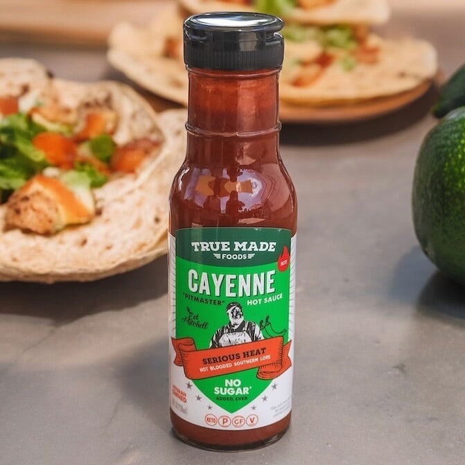 Bottle of True Made Foods Cayenne Hot Sauce on a table with tacos in the background.