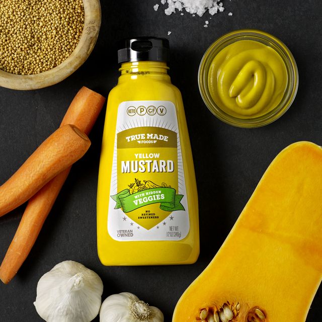True Made Foods Yellow Mustard bottle with garlic, butternut squash, carrots, and other vegetables surrounding it.